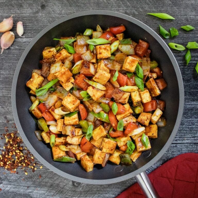 Skillet stir-fry with tofu cubes, bell peppers, and onions garnished with green onions on a dark pan.