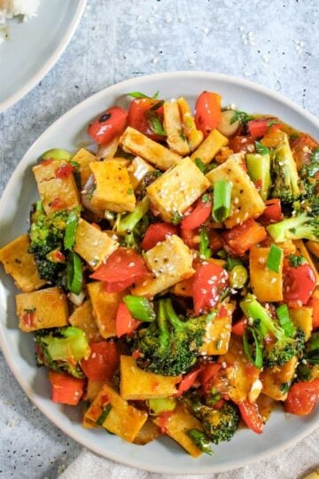 Stir-fry plate with cubed tofu, broccoli, bell peppers, tomatoes, and sesame seeds on a light blue plate