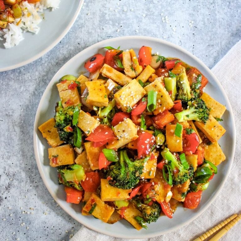 Stir-fry plate with cubed tofu, broccoli, bell peppers, tomatoes, and sesame seeds on a light blue plate