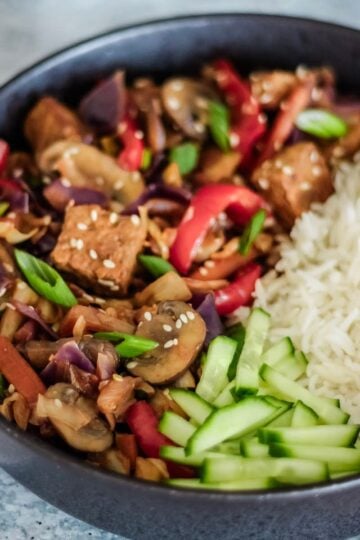 Bowl of tofu and vegetable stir-fry with sesame seeds, cucumber slices, and white rice on the side.
