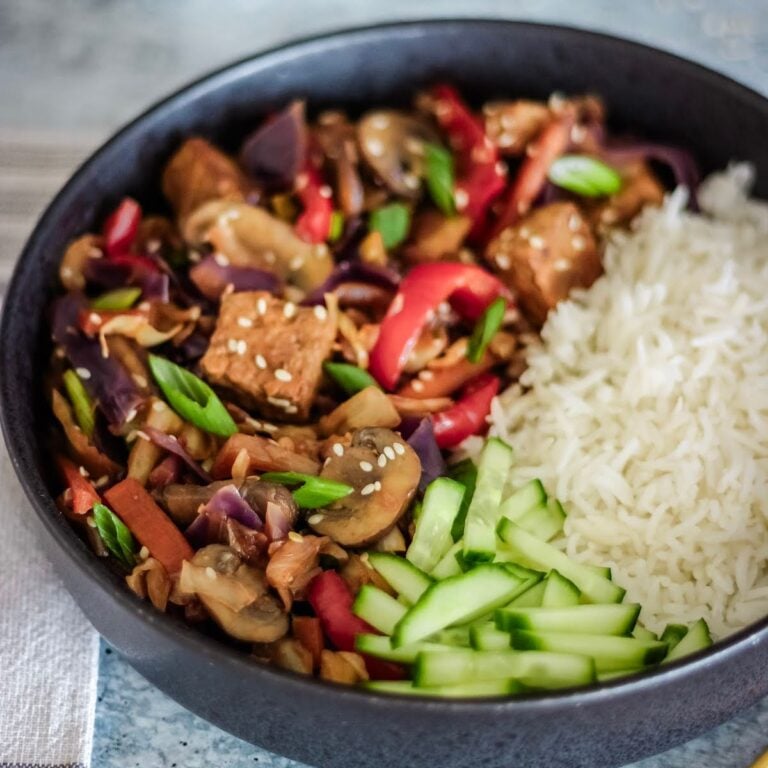 Bowl of tofu and vegetable stir-fry with sesame seeds, cucumber slices, and white rice on the side.