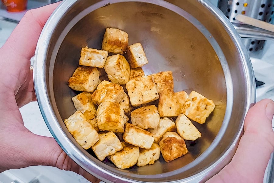 Bite-sized, pan-fried tempeh cubes with golden-brown crust in a stainless steel bowl being held by two hands in a kitchen.