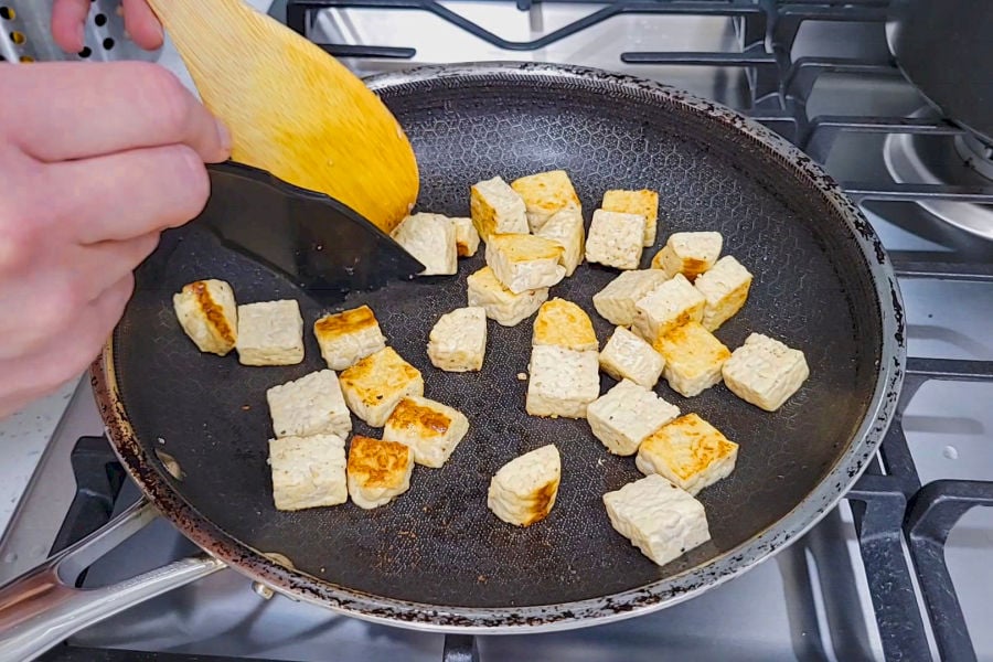 Tempeh cubes browning in a frying pan, being turned with a yellow spatula over a gas burner