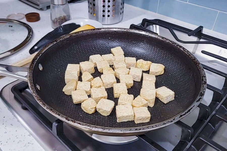Cubed tempeh browning in a large nonstick skillet on a gas stove.