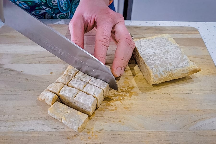 Person slices a block of tempeh on a wooden cutting board, cutting it into cubes.